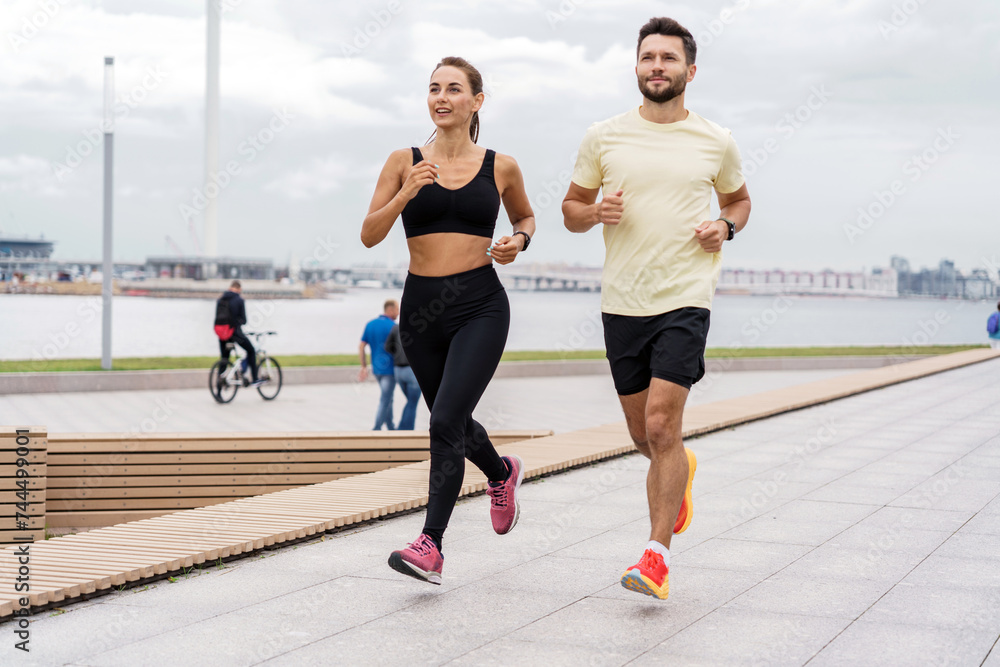 Couple in sportswear runs together on a city promenade with a marine backdrop.