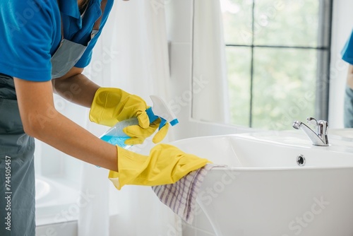 Focused on chores a woman in uniform sprays and cleans bathroom sink and faucet. Her dedication to housework emphasizes purity hygiene and shining fixtures at home. spray cleaner photo