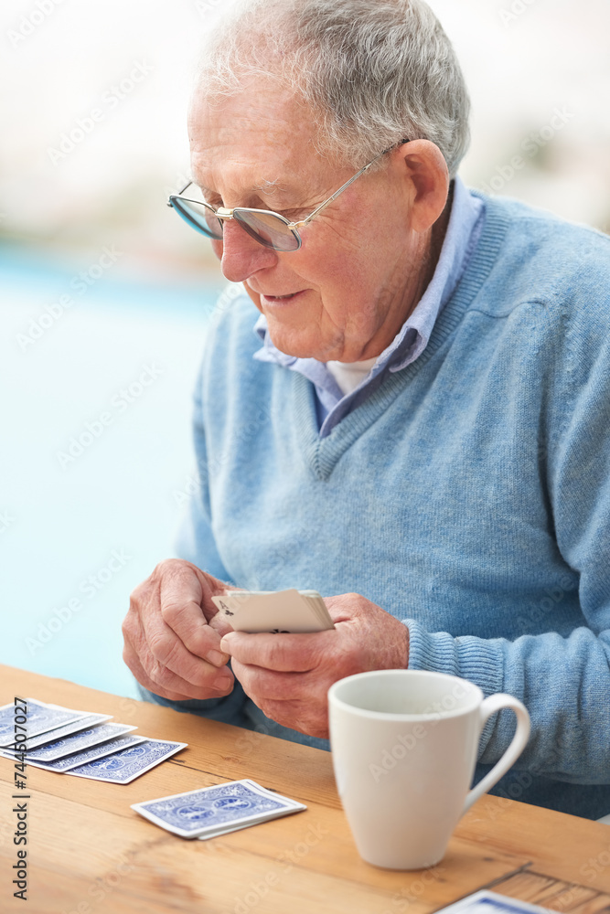 Elderly man, coffee and playing cards on table for poker, relax and happy in retirement. Senior person, grandfather and beverage with thinking, hobby and fun strategy on patio in home drinking tea