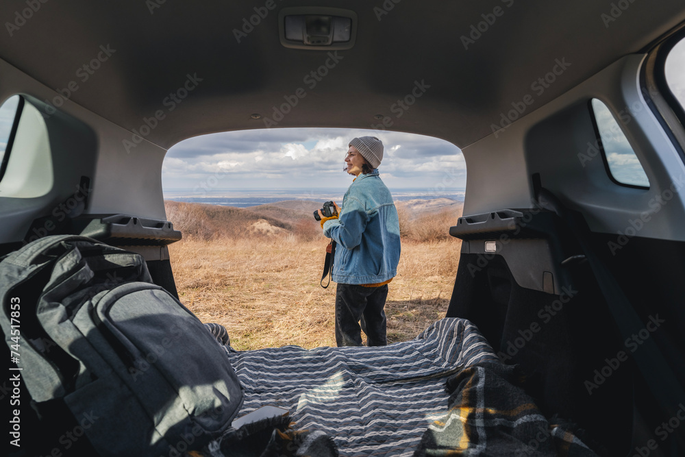 Young happy woman sitting in an open car trunk taking pictures with a camera. Travel by car