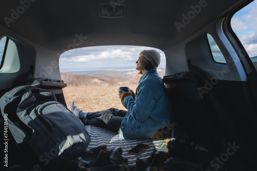 A young happy woman sitting in an open car trunk drinking tea from a mug. Travel by car concept