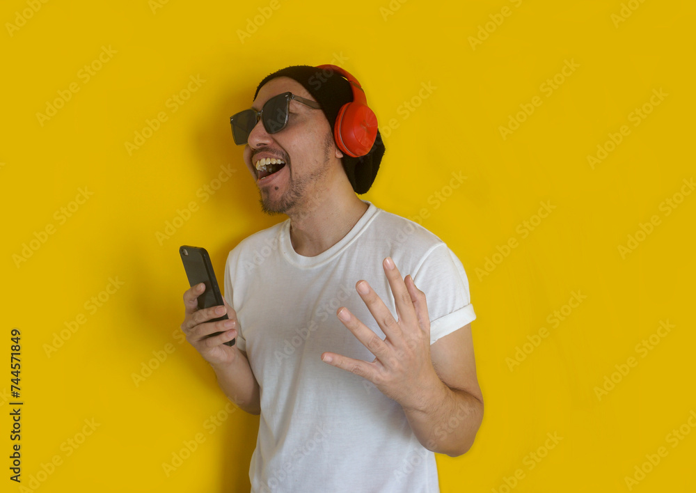 Young man singing his favorite song, using smartphone as microphone, wearing sunglasses, beanie hat and white t shirt, isolated on yellow background Young man singing his favorite song, using smartphone as microphone, wearing sunglasses, beanie hat and white t shirt, isolated on yellow background