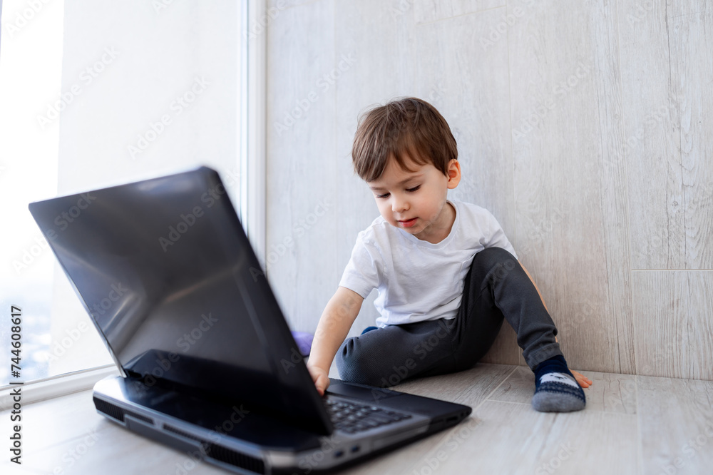 Little boy sits on the floor and plays on laptop. The child is sitting in front of window. Little boy sits on the floor and plays on laptop. The child is sitting in front of window.