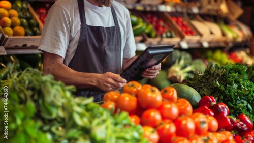 Grocery store worker with apron using tablet among fresh vegetables.