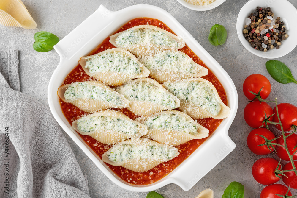 Raw conchiglioni pasta stuffed ricotta cheese and spinach with tomato sauce and parmesan cheese on top in a white baking dish on a gray concrete background.
