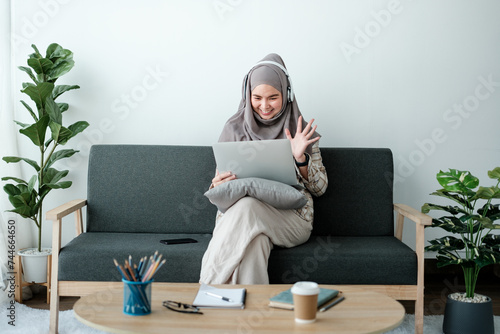 Beautiful asian woman in hijab works inside a modern office building, Muslim talks on video call, online meeting with colleagues and clients, woman cheerfully waves her hand and greets interlocutors photo
