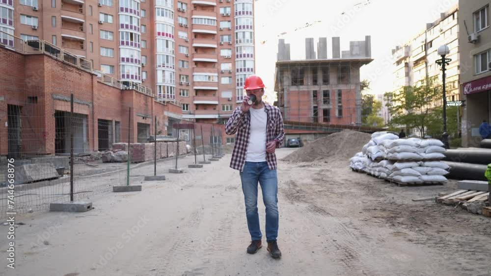 Construction engineer walking through construction site and drinking coffee. Builder in hard hat, checkered shirt and jeans walks along a building sites and has cup of coffee. Architect having break. Construction engineer walking through construction site and drinking coffee. Builder in hard hat, checkered shirt and jeans walks along a building sites and has cup of coffee. Architect having break.