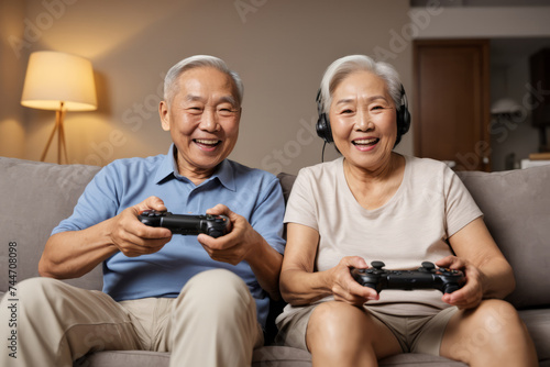 Elderly smiling Asian couple holding gaming controllers and playing videogames on the living room couch at home at night, aged gamer man and woman enjoying active life after retirement