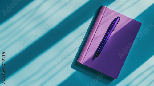 a purple notebook with a pen on top of it next to a shadow of a sheet of paper on a blue background. photo