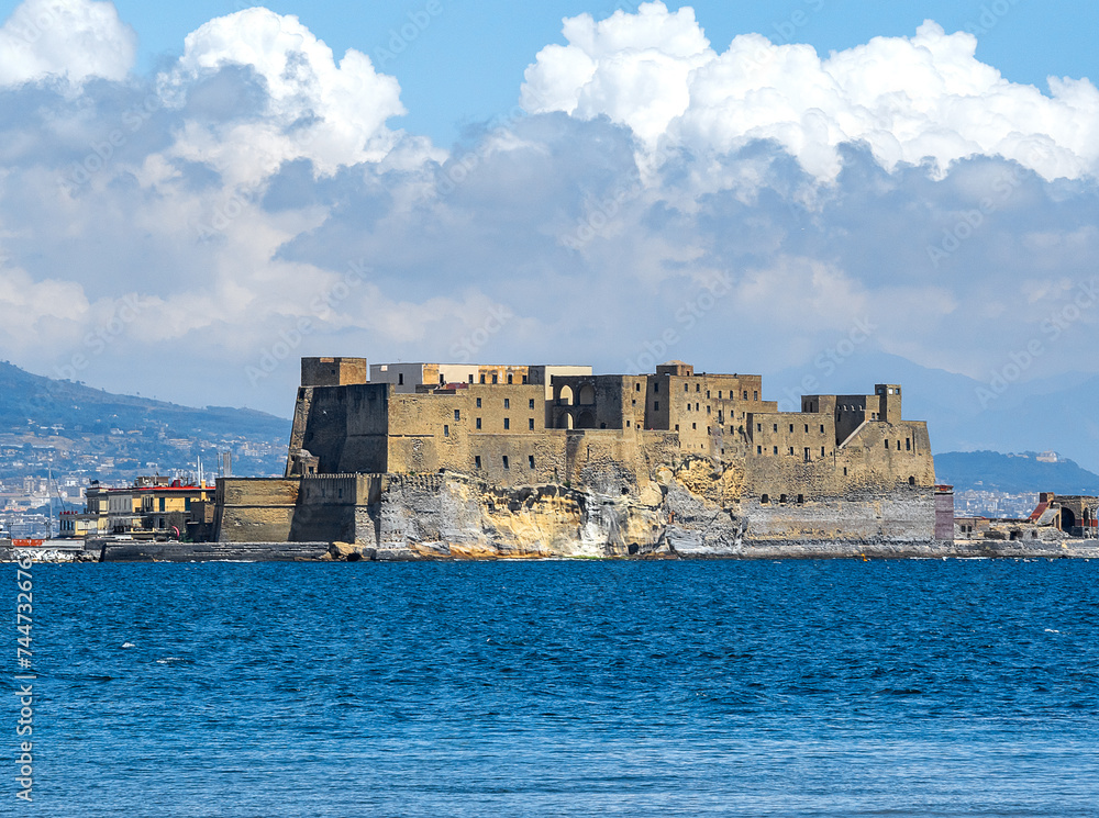 On the Islet of Megaride emerging from the deep blue sea is one of the oldest castles in Naples, Castel dell'Ovo, a fortress of Norman origin. Blue sky with beautiful white clouds on a sunny day.
