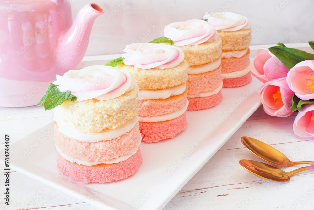 Spring mini cakes with buttercream rose. Table scene with a white wood background. Pink layers with flower topping. Spring mini cakes with buttercream rose. Table scene with a white wood background. Pink layers with flower topping.