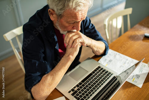 Senior man contemplating while looking at laptop with paperwork photo