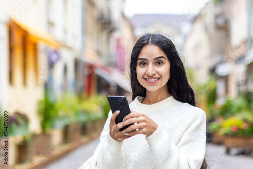 Portrait of a smiling and beautiful Indian girl standing in the middle of a city street, holding a mobile phone and looking at the camera