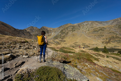 Person in middle of Pyrenees mountain hiking with walking sticks crossing bridge and lake in background