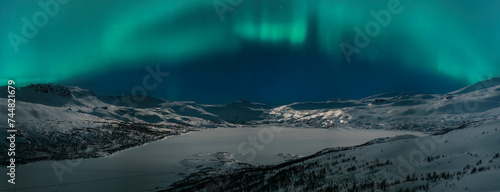 Wide scenic night panorama of winter mountains highlighted by Aurora Borealis green lights. Norway Swedish Northern mountains area