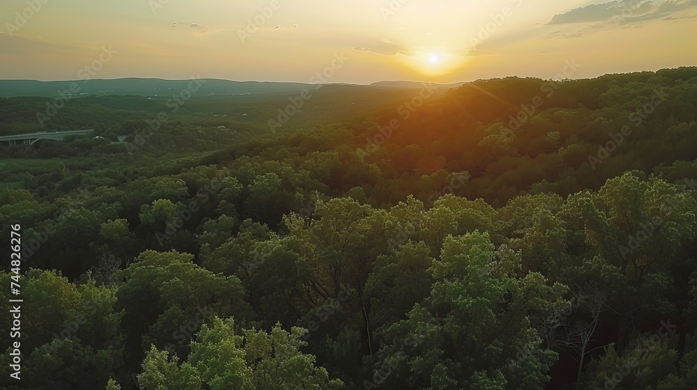 A bird's-eye view reveals a breathtaking sunset casting its warm, dramatic colors over forested hills, creating a scene of serene beauty and natural splendor.