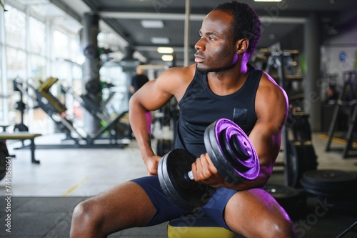 Young African American man sitting and lifting a dumbbell close to the rack at gym
