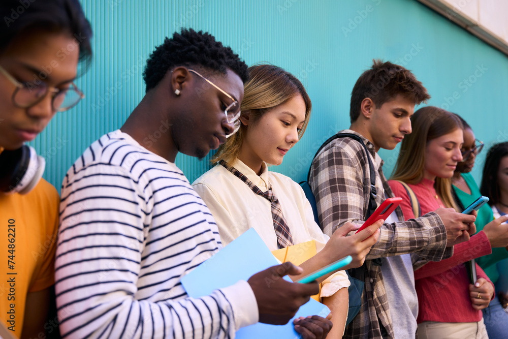 Obraz premium Serious international students using smart phone. Young people multicultural friends concentrated in a mobile cell. Technology addicted group of people in a blue wall background