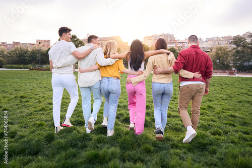 Rear view of a Group of young people walking towards the sunset in the grass of the park. They are hugging each other enjoying their friendship. Students carefree enjoying freedom and youth together