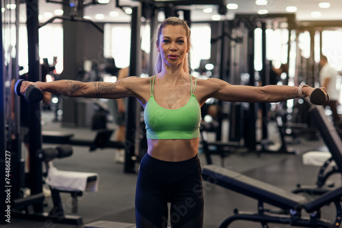 Muscular fitness woman athlete doing heavy weight exercise for shoulders with dumbbells in the gym