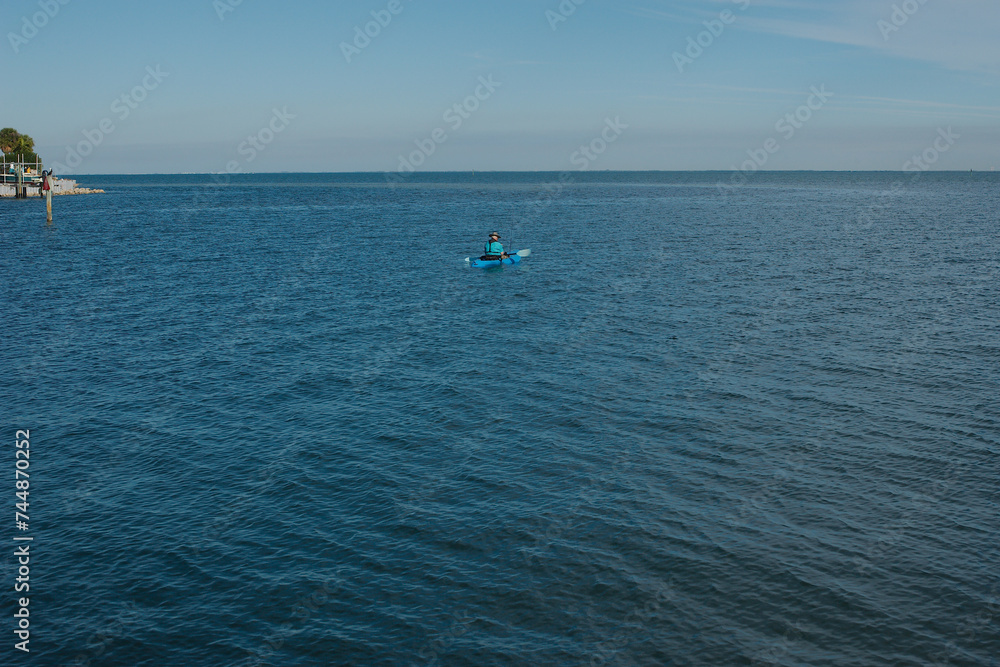 Man in blue kayak in the middle with wood post and seawall on the left looking out to Tampa Bay and blue calm water on a sunny afternoon. Horizontal shot with room for copy.  blue sky in the back. 