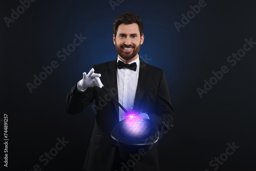 Magician showing trick with wand and top hat on dark background. Fantastic light coming out of hat