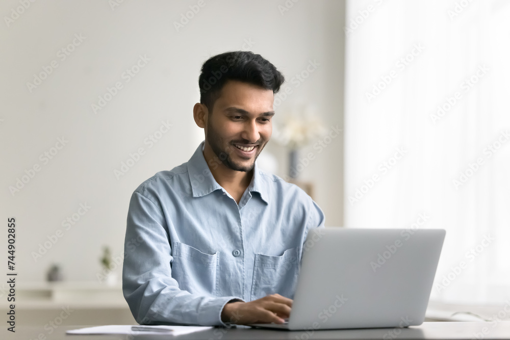 Smiling young Indian man working or studying using wireless computer, typing text on laptop keyboard, share messages, solve business by e-mailing to client, read good news. Modern tech usage, workflow Smiling young Indian man working or studying using wireless computer, typing text on laptop keyboard, share messages, solve business by e-mailing to client, read good news. Modern tech usage, workflow