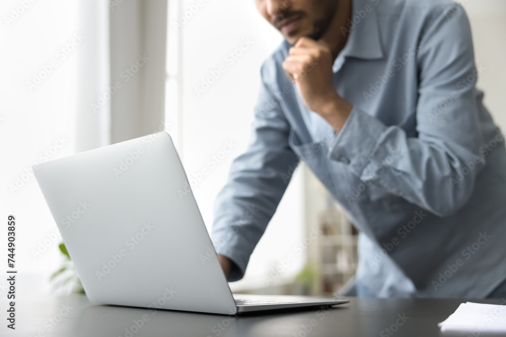Cropped close up of pensive Indian man worker, freelancer doing task leaned at table with laptop, search issue solution, looks pensive and concentrated, pondering ideas, engaged in on-line workflow Cropped close up of pensive Indian man worker, freelancer doing task leaned at table with laptop, search issue solution, looks pensive and concentrated, pondering ideas, engaged in on-line workflow