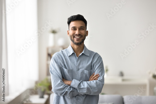 Portrait of happy Indian man having wide toothy smile, pose with arms-crossed, look at camera, spend carefree time in modern living room, enjoy day-off. Profile picture of homeowner, renter or tenant photo