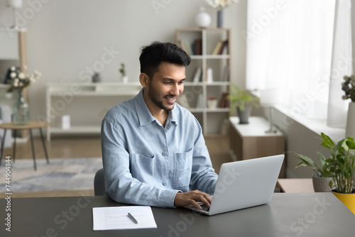 Indian businessman, freelance worker sit at desk with computer, working on-line from home, using laptop, typing message, solve business by e-mailing, correspond to client, do task. Workflow using tech photo