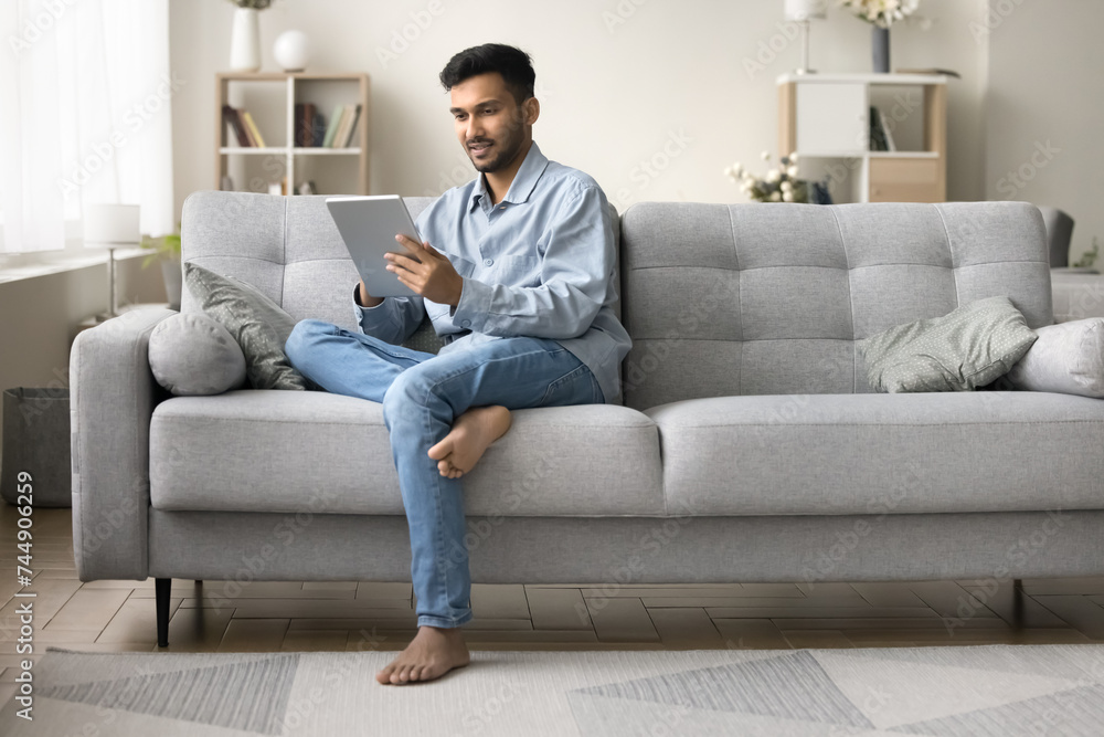 Indian man rest on couch in living room look at digital tablet screen, read e-mail, chatting, browsing wireless internet, text message on pad gadget, working on-line, buying goods via e-commerce app