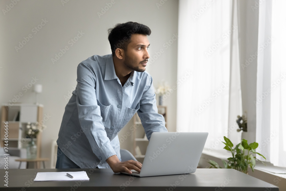 Thinking process of young Indian freelance worker standing leaned at workplace desk with laptop, ponders, looking out window, deep in thoughts, considering strategy, planning start up, work on project Thinking process of young Indian freelance worker standing leaned at workplace desk with laptop, ponders, looking out window, deep in thoughts, considering strategy, planning start up, work on project