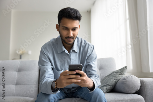 Serious engrossed Indian guy read sms, lead on-line business or personal chat sit on sofa at home, learn e-mail commercial offer, using new mobile application, watching video spend pastime on internet