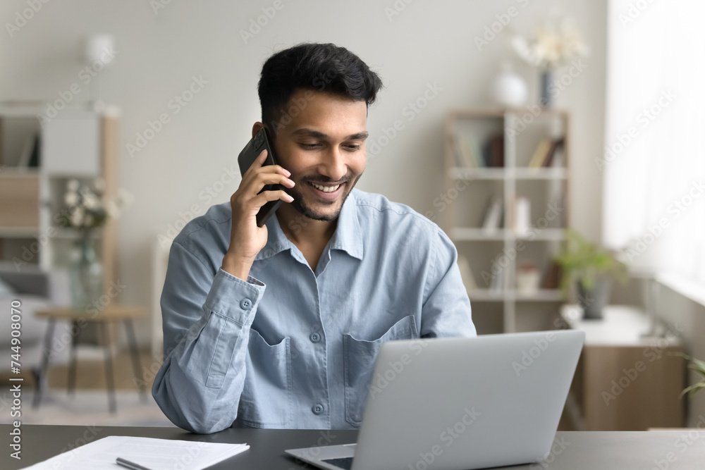 Smiling Indian guy provide information to client by phonecall, sit at desk look at laptop screen, explain offer detail, buy, order remotely, talk to customer service, receive help. Tech, communication Smiling Indian guy provide information to client by phonecall, sit at desk look at laptop screen, explain offer detail, buy, order remotely, talk to customer service, receive help. Tech, communication