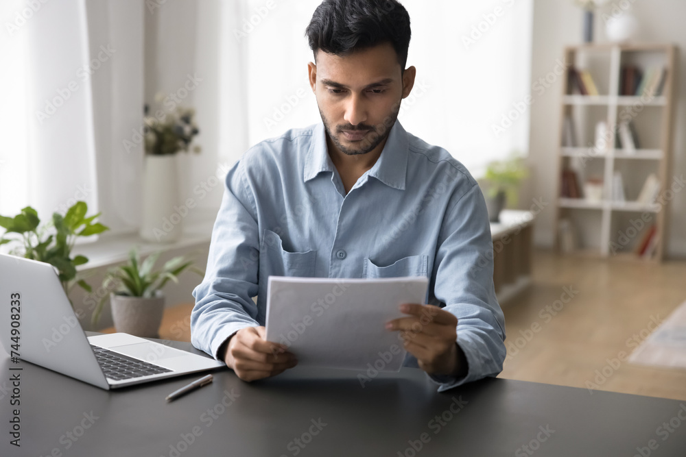 Serious Indian business man working with papers, holding official letter, read document think on conditions, learning terms, engaged in paperwork, prepare text of agreement at homeoffice with laptop Serious Indian business man working with papers, holding official letter, read document think on conditions, learning terms, engaged in paperwork, prepare text of agreement at homeoffice with laptop