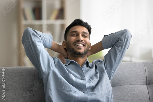 Satisfied Indian guy enjoy pleasant relaxation alone indoor, leaned on comfortable sofa, put hands behind head, spend weekend in cozy living room, breathing fresh conditioned air in modern smart home photo