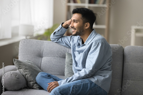 Satisfied handsome Indian guy relaxing seated on cozy sofa smiling looking out window admire view, enjoy pleasant daydreams on carefree weekend at own or rented apartment. Accommodation, day-off, rest © fizkes