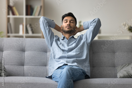 Indian man fell asleep leaned on couch relaxing with eyes closed, put hands behind head breath fresh air in summer day inside living room with climate control. Rest, carefree day-off, fatigue relieve photo