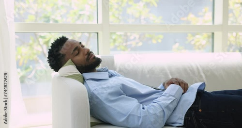 Peaceful sleepy handsome young African man resting on comfortable couch, lying with closed eyes, breathing fresh cool air, enjoying leisure, relaxation, work break at cozy home photo