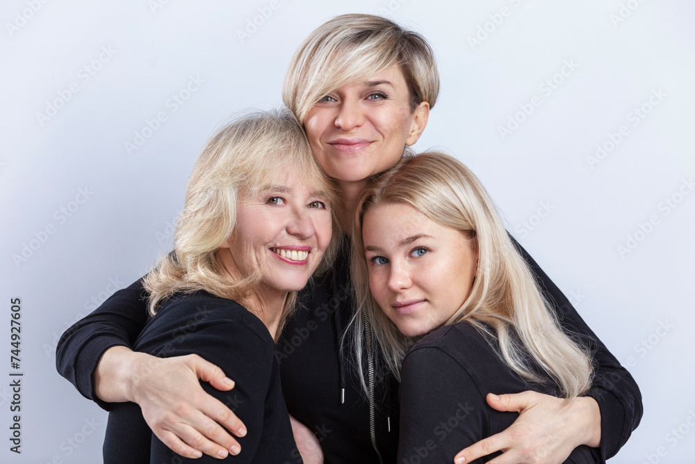 Three generations of women hug and smile. Beautiful blondes in black sweaters. Love and tenderness. Light background.