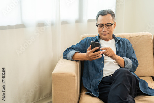 Fototapeta Naklejka Na Ścianę i Meble -  Handsome asian mature old man using smartphone on sofa in living room at home. Happy Portrait of cheerful smiling senior asian man holding cell phone. Mature People and lifestyle