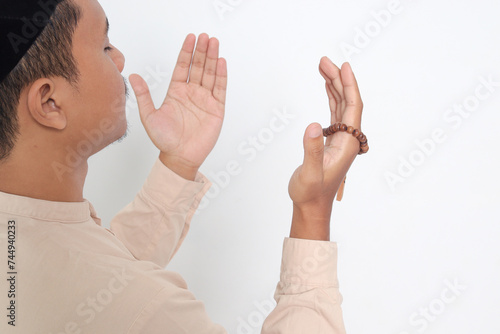 Back view portrait of religious Asian muslim man in koko shirt with skullcap praying earnestly with his hands raised, holding islamic beads. Devout faith concept. Isolated image on white background