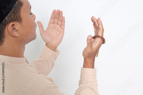 Back view portrait of religious Asian muslim man in koko shirt with skullcap praying earnestly with his hands raised, holding islamic beads. Devout faith concept. Isolated image on white background photo