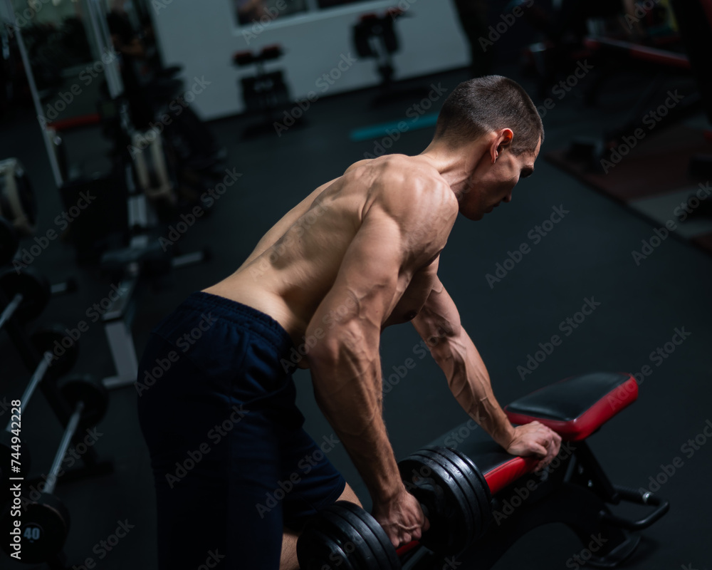 Shirtless man doing dumbbell row to waist on bench at gym. 