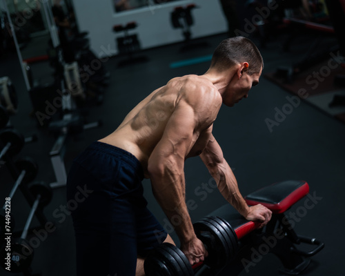 Shirtless man doing dumbbell row to waist on bench at gym. 