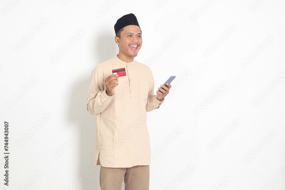 Portrait of attractive Asian muslim man in koko shirt with skullcap holding a mobile phone and presenting credit card. Isolated image on white background Portrait of attractive Asian muslim man in koko shirt with skullcap holding a mobile phone and presenting credit card. Isolated image on white background