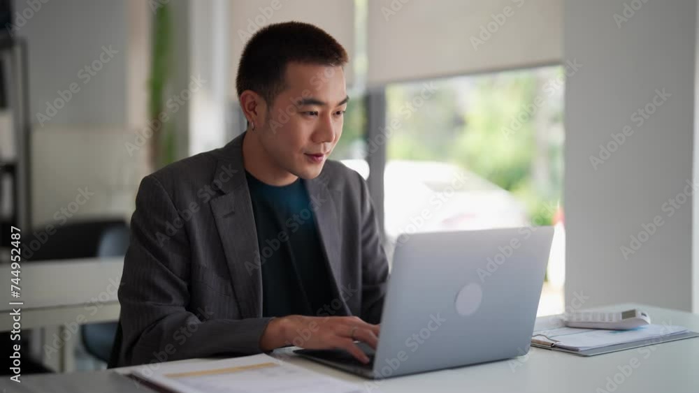 Young Asian businessman typing work information on his laptop at the work desk Young Asian businessman typing work information on his laptop at the work desk
