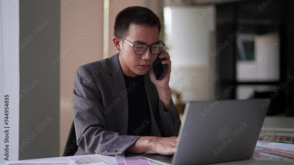 Attentive Asian interior designer multitasking, having a focused conversation on the phone and working on his laptop at the work desk Attentive Asian interior designer multitasking, having a focused conversation on the phone and working on his laptop at the work desk