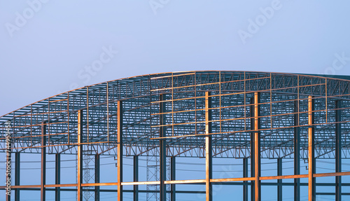 Large metal industrial factory building with dome roof outline structure in construction site against blue evening sky background at sunset time