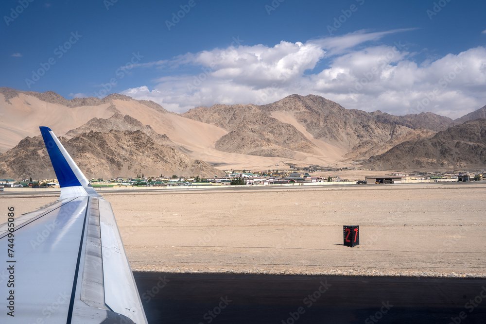 A shot from airplane window, visible plane wings. Dry Mountains with runway in the foreground and mountains in the back with clear blue sky and clouds